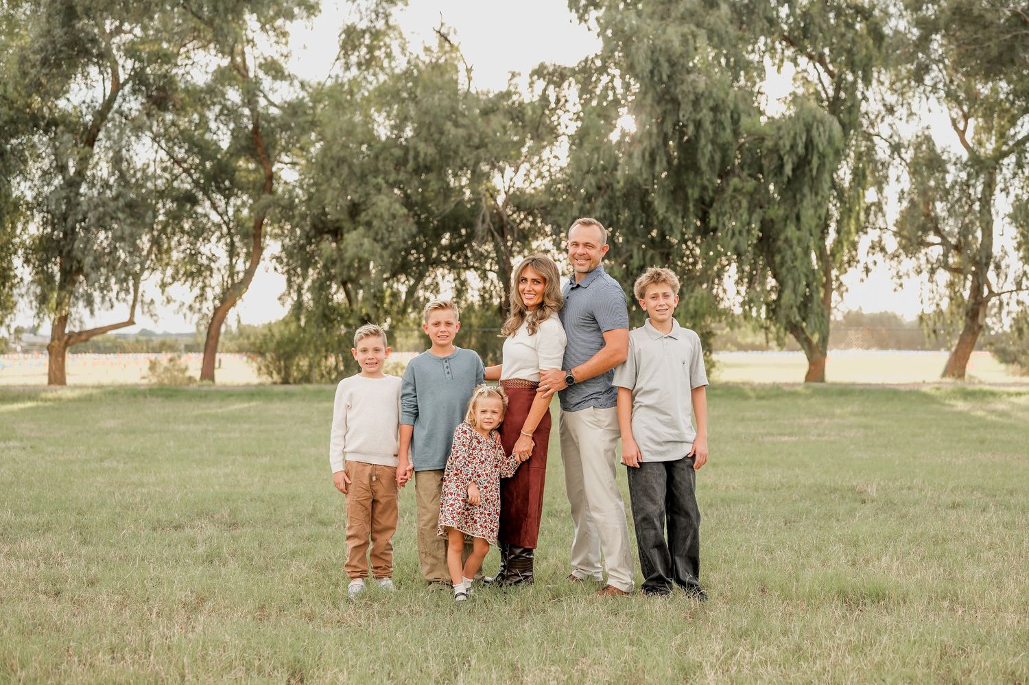 Family of six posing outdoors in a green field with trees, wearing coordinated casual outfits for a professional photo.