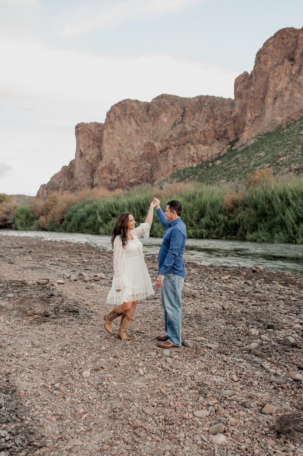 Couple dancing on rocky riverbank with red rock mountains and lush greenery in the background.