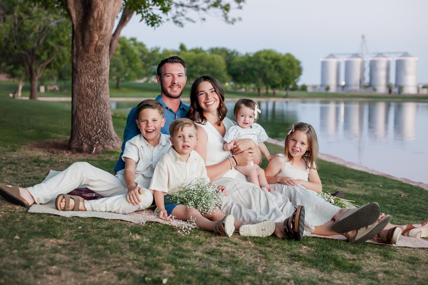 Happy family of seven smiling together on a grassy lakeside park, with grain silos visible in the background.