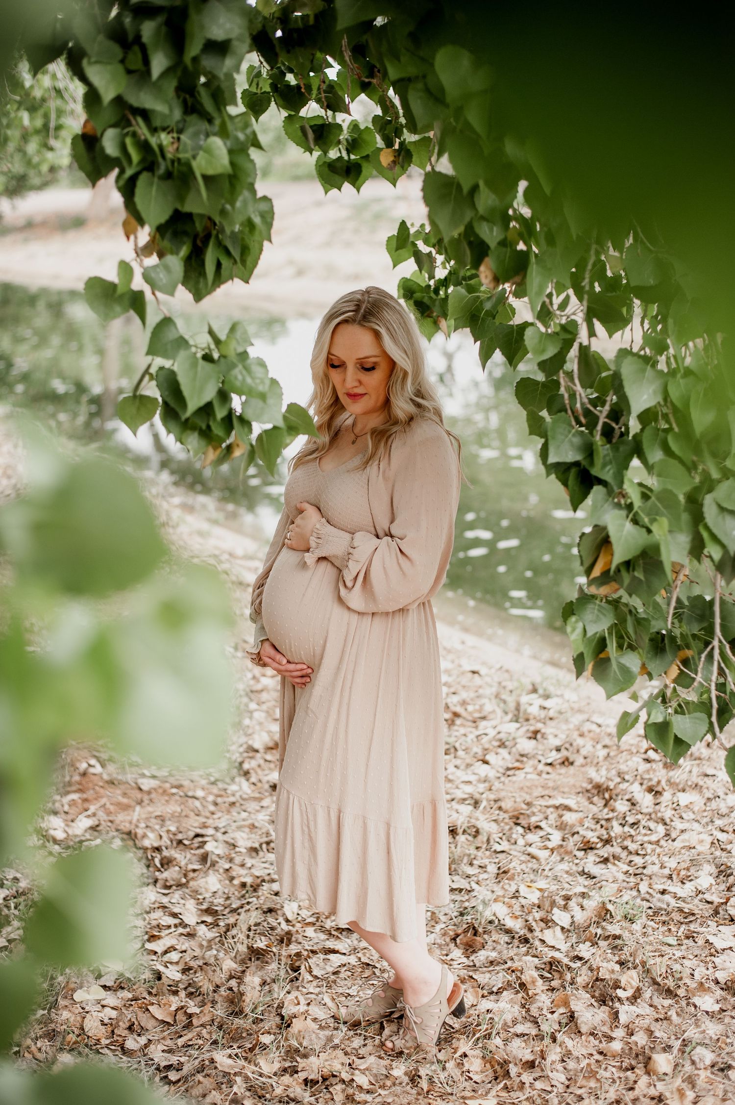 A woman in a flowing beige dress poses thoughtfully in a natural outdoor setting with green foliage and fallen leaves.