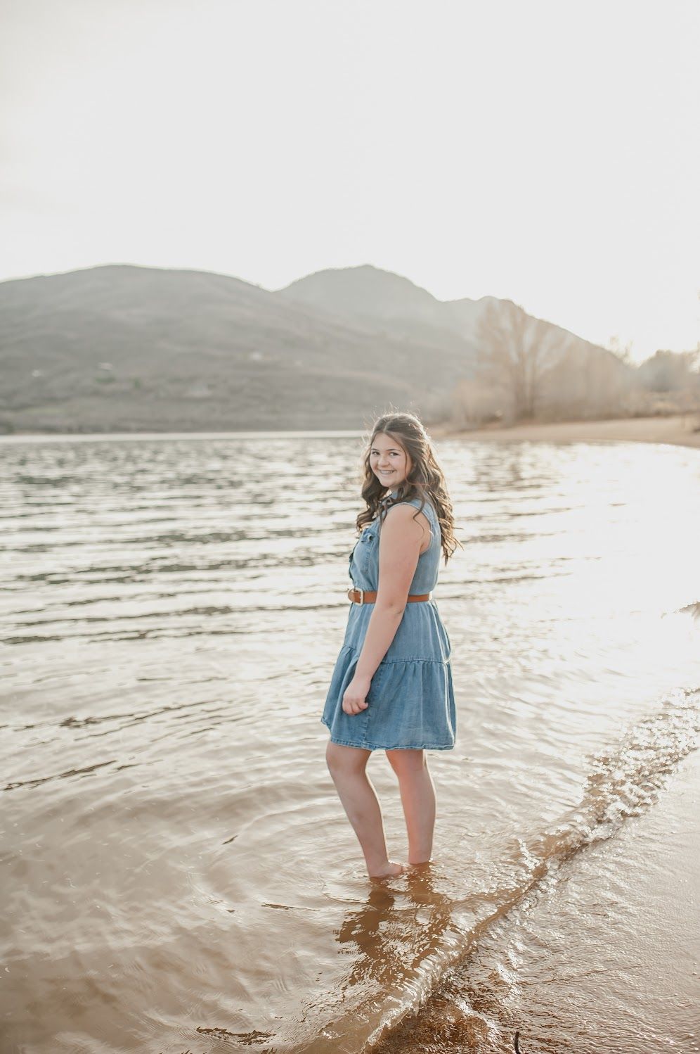 Young woman in blue denim dress standing in shallow lake water with mountain backdrop in warm sunlight.