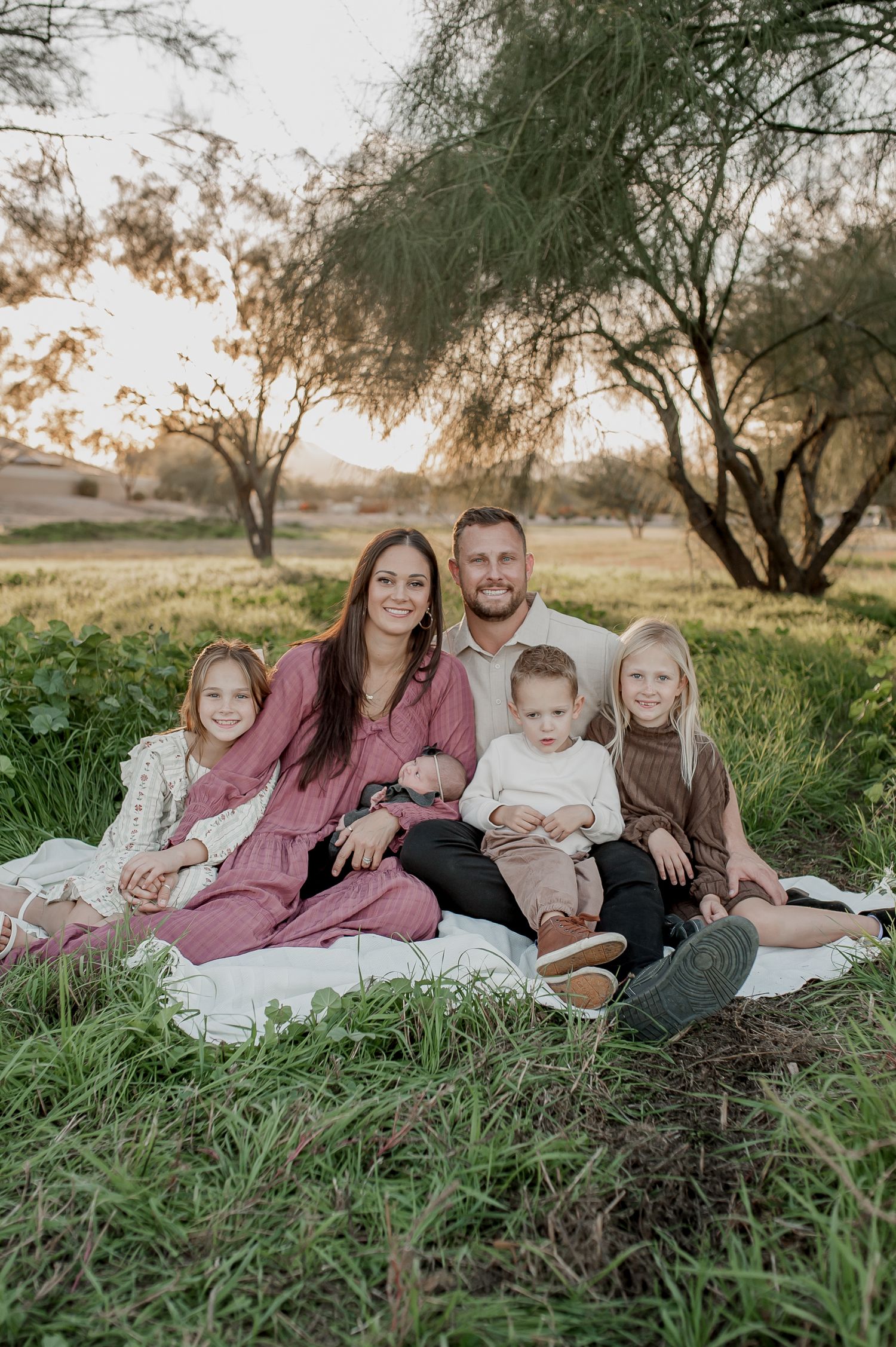 Family of five sitting on a blanket in a grassy field with trees at golden hour.