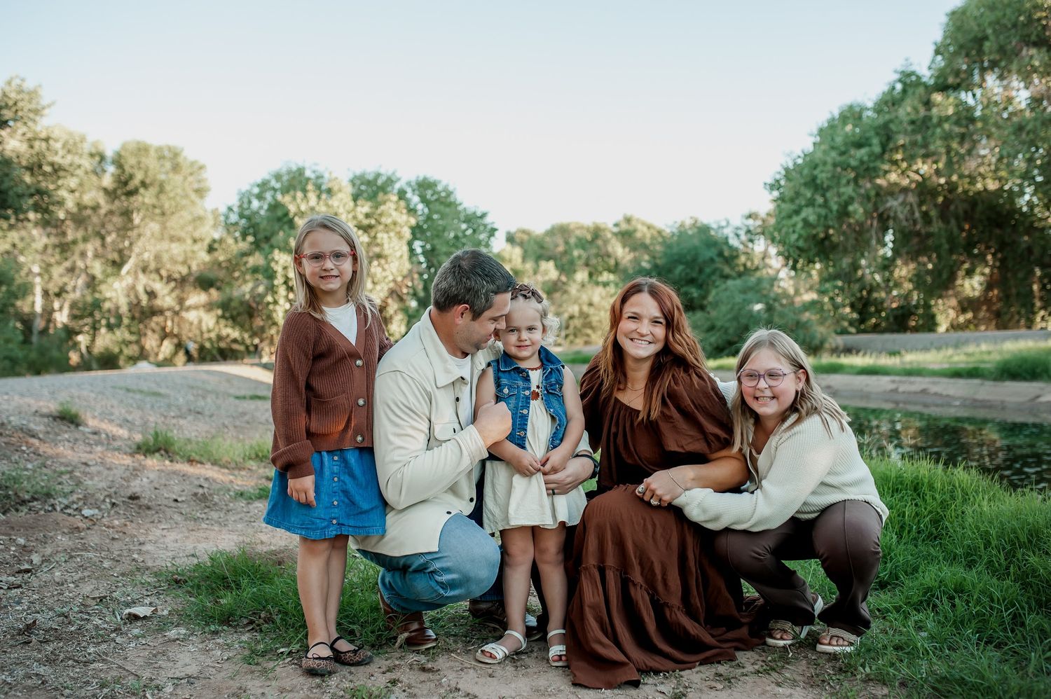 Family of five posing outdoors on a dirt path surrounded by green trees during golden hour.