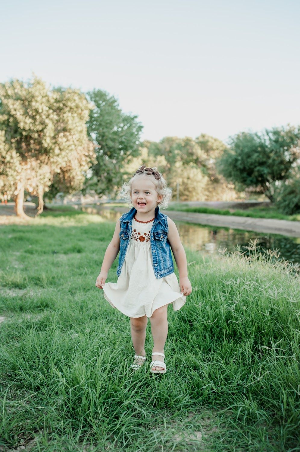 A cheerful toddler girl in a white dress and denim vest plays on green grass near a pond in a sunny park.