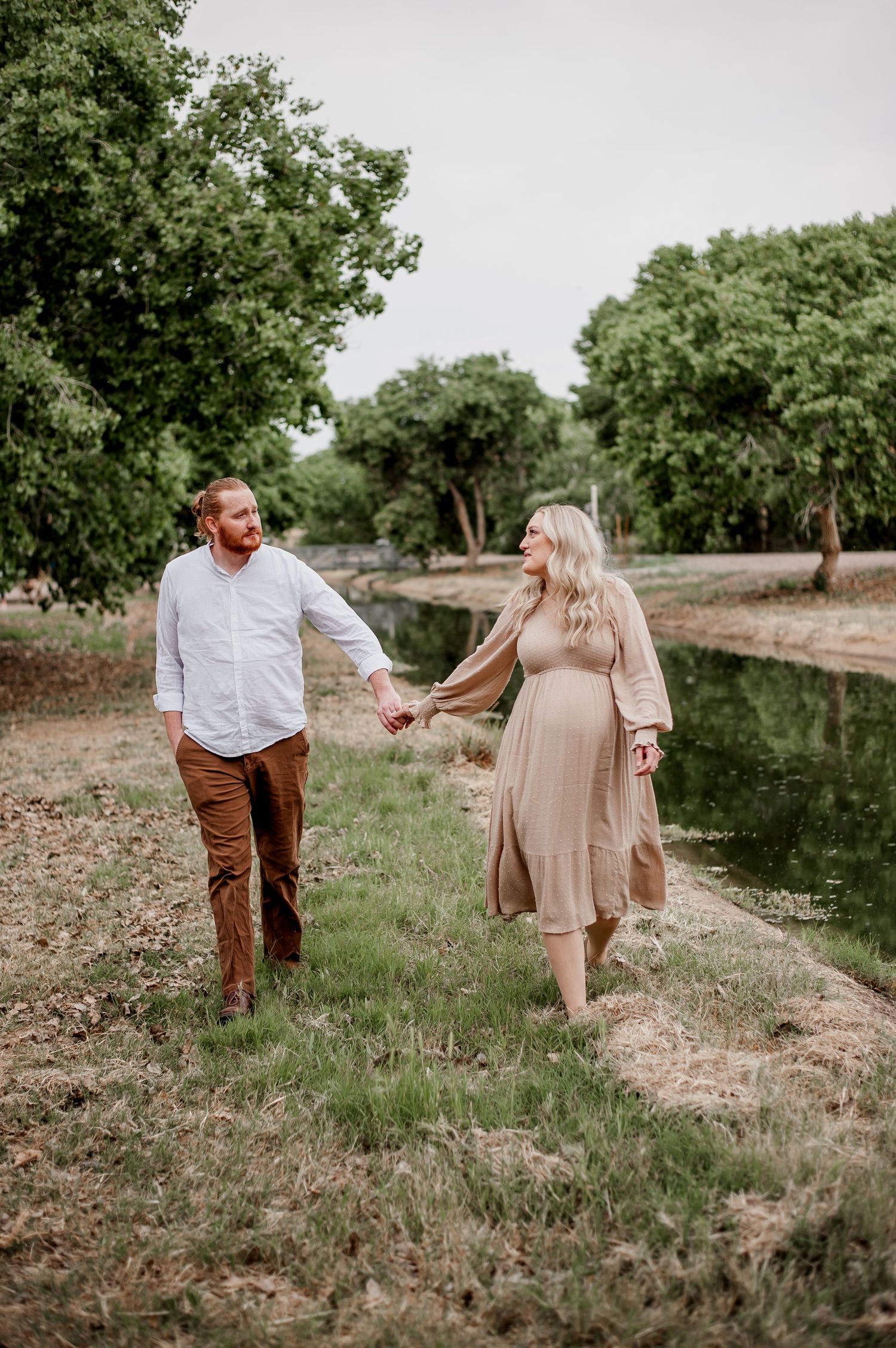 A couple walks together on a grassy hillside path near a pond, surrounded by trees on a cloudy day.