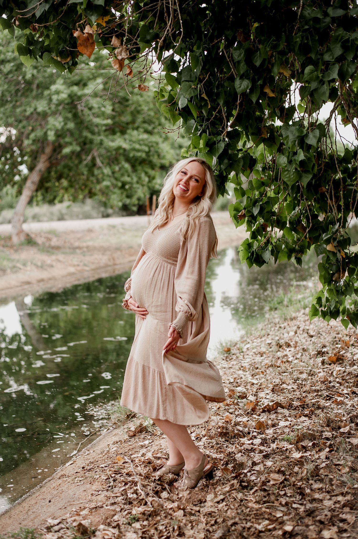 A pregnant woman in a flowing beige dress poses gracefully by a tranquil pond surrounded by greenery.