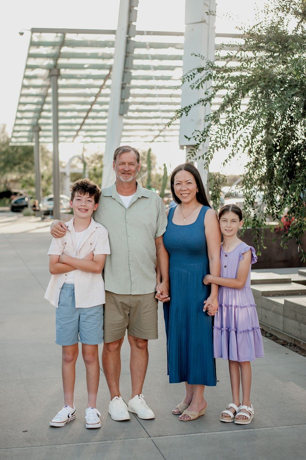 Family of four smiling outdoors under a pergola, parents with two children in casual summer attire.