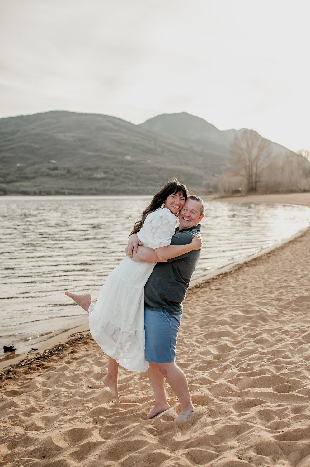 Couple embracing on sandy beach shore with mountain lake backdrop, man lifting woman in white dress.