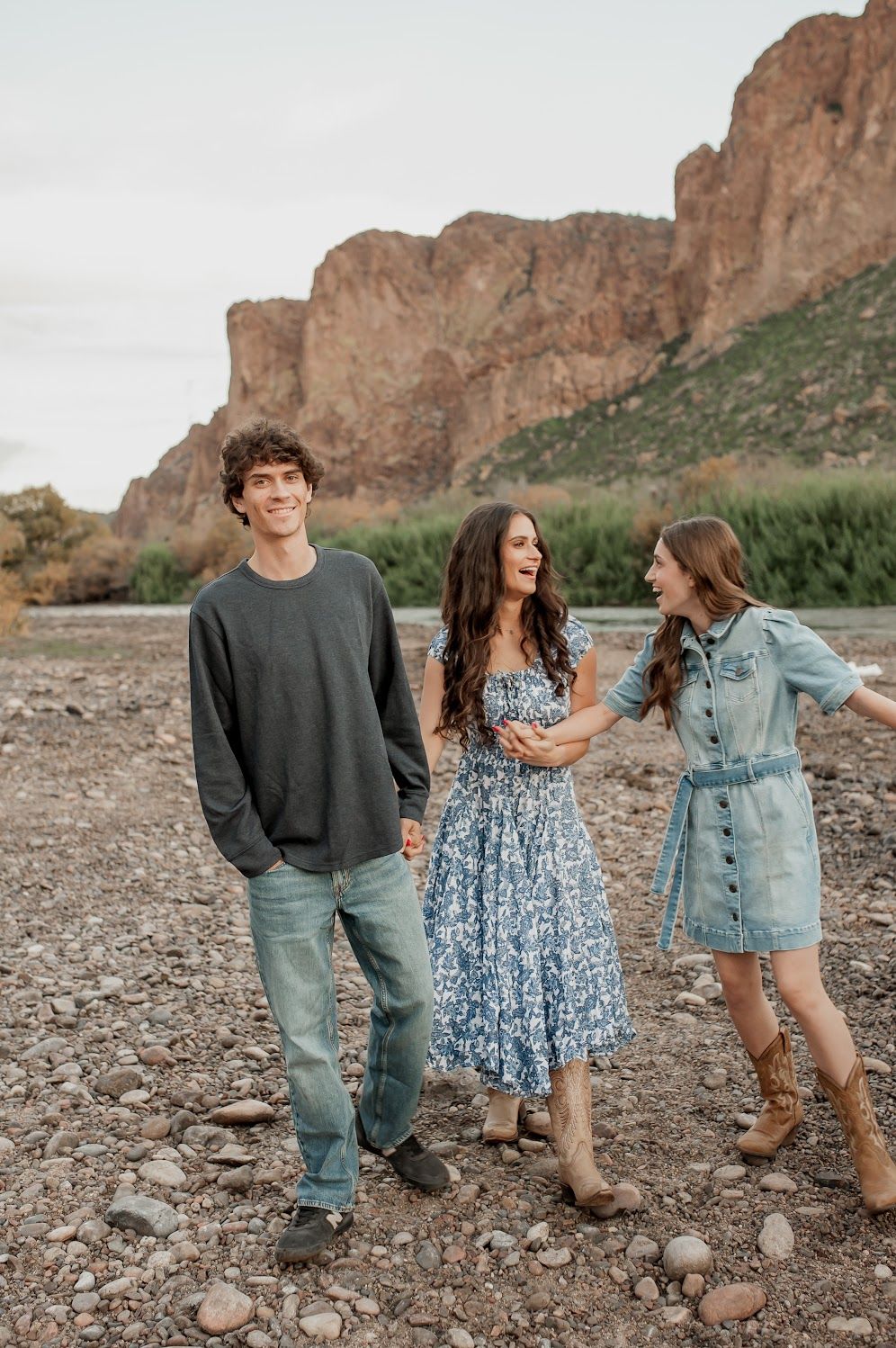 Three young people smiling and laughing outdoors on a rocky riverbed with dramatic desert cliffs in the background.
