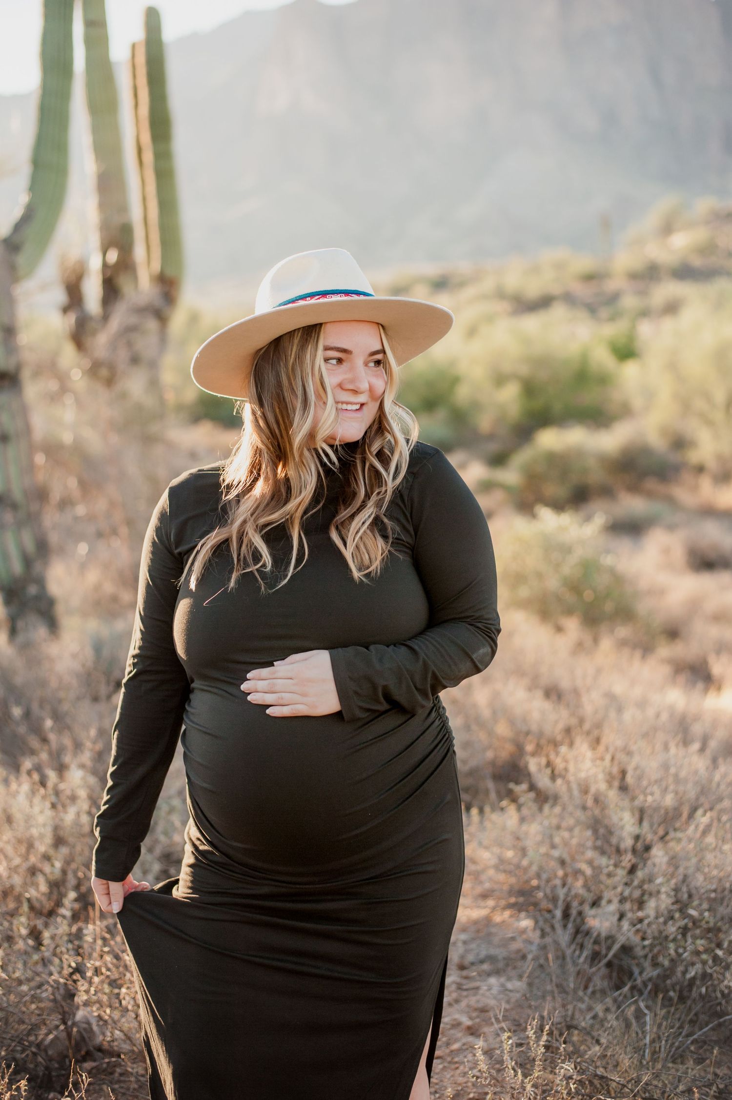 A series of desert maternity photos showing a woman in a black dress and tan hat posing among cacti at sunset.