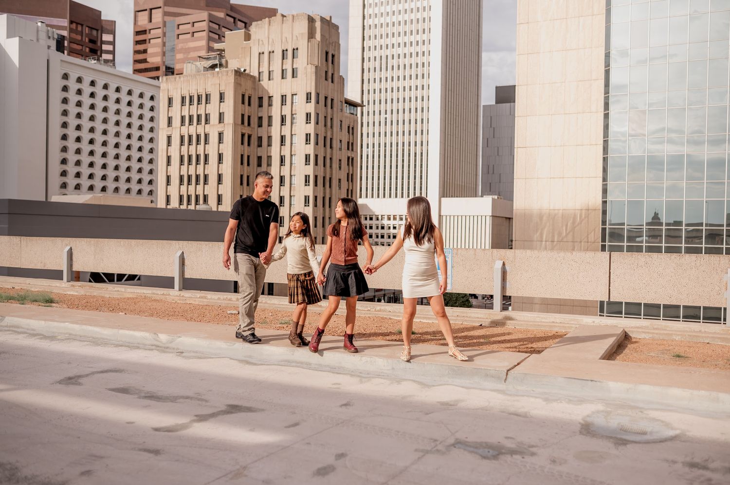 Family of four holding hands and walking along a city sidewalk with tall downtown buildings in the background.