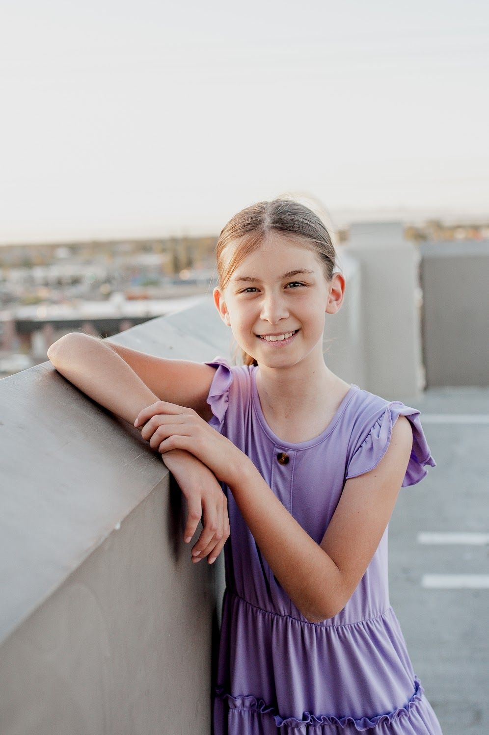 A smiling young girl in a purple ruffle dress leans on a concrete ledge outdoors, posing for a portrait photo.