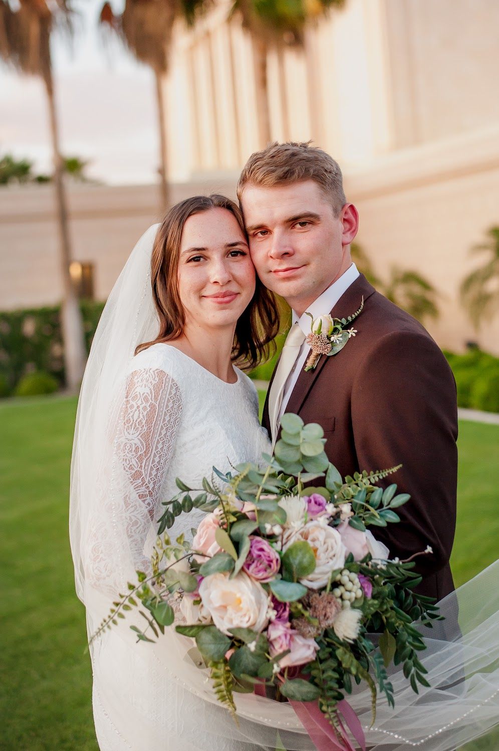 Bride and groom pose together outdoors, she holds a pink and white bouquet, wearing lace dress and veil, he wears brown suit.