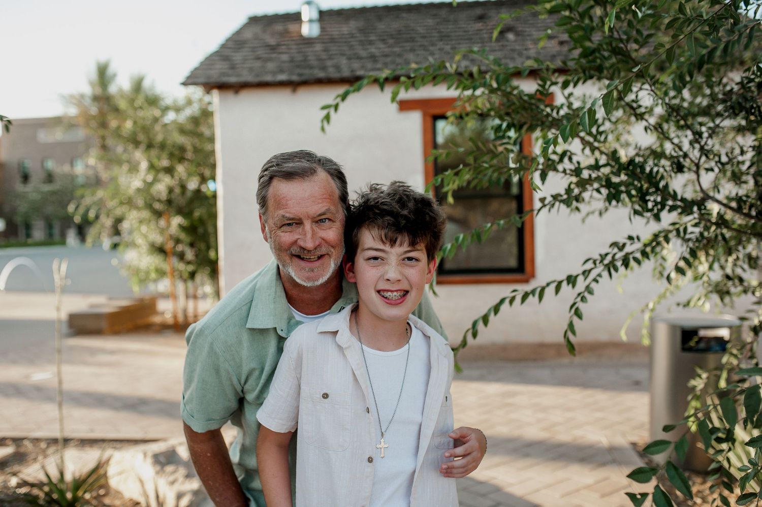 Smiling grandfather and grandson posing together outdoors in front of a white house surrounded by green trees.