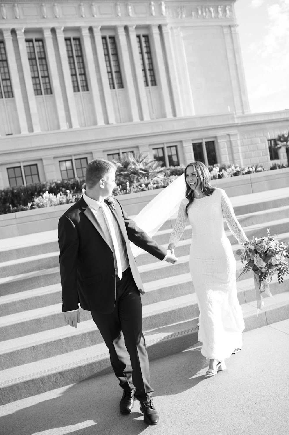 Black and white photo of a bride and groom holding hands walking up steps outside a grand building on their wedding day.