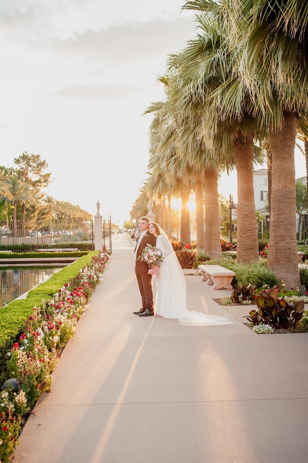 Bride and groom pose on a palm-lined walkway at golden hour, surrounded by colorful flowers at a tropical wedding venue.