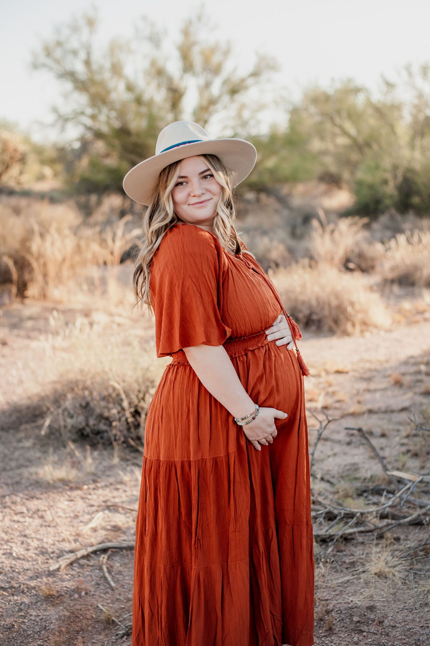 A woman in a flowing rust-colored dress and beige hat stands in a desert setting with dried brush surrounding her.