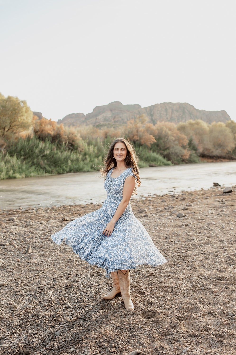 Young woman in blue floral dress twirling by a river with rocky mountains and trees in the background.