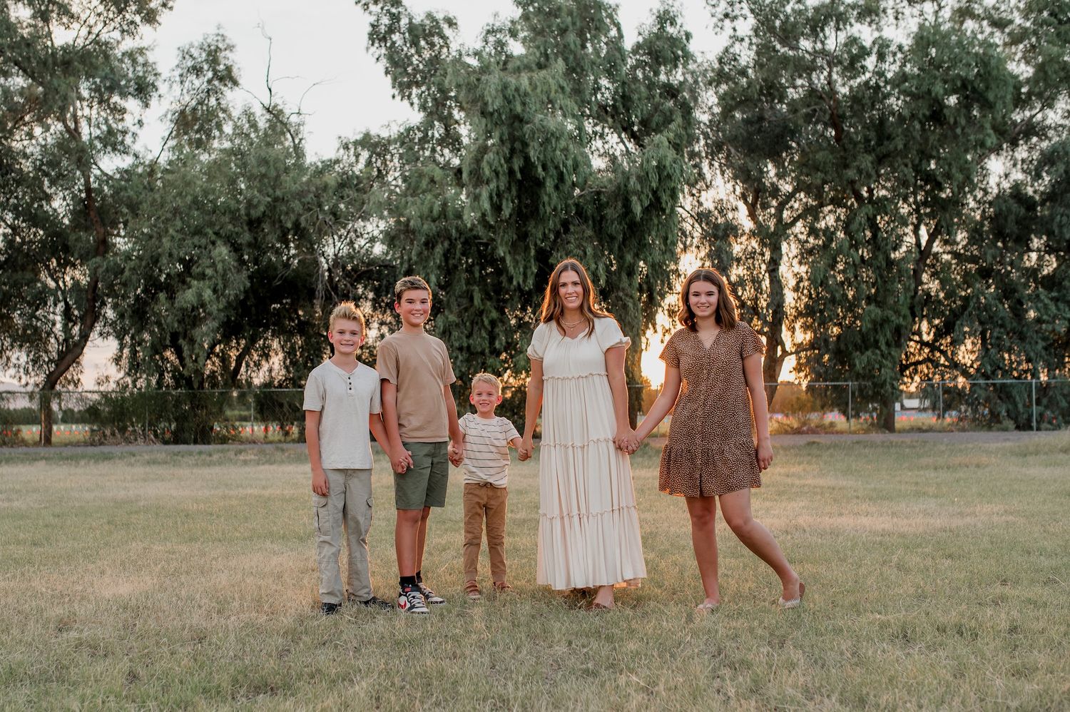 Family of five posing outdoors at golden hour in a grassy field with green trees in the background.