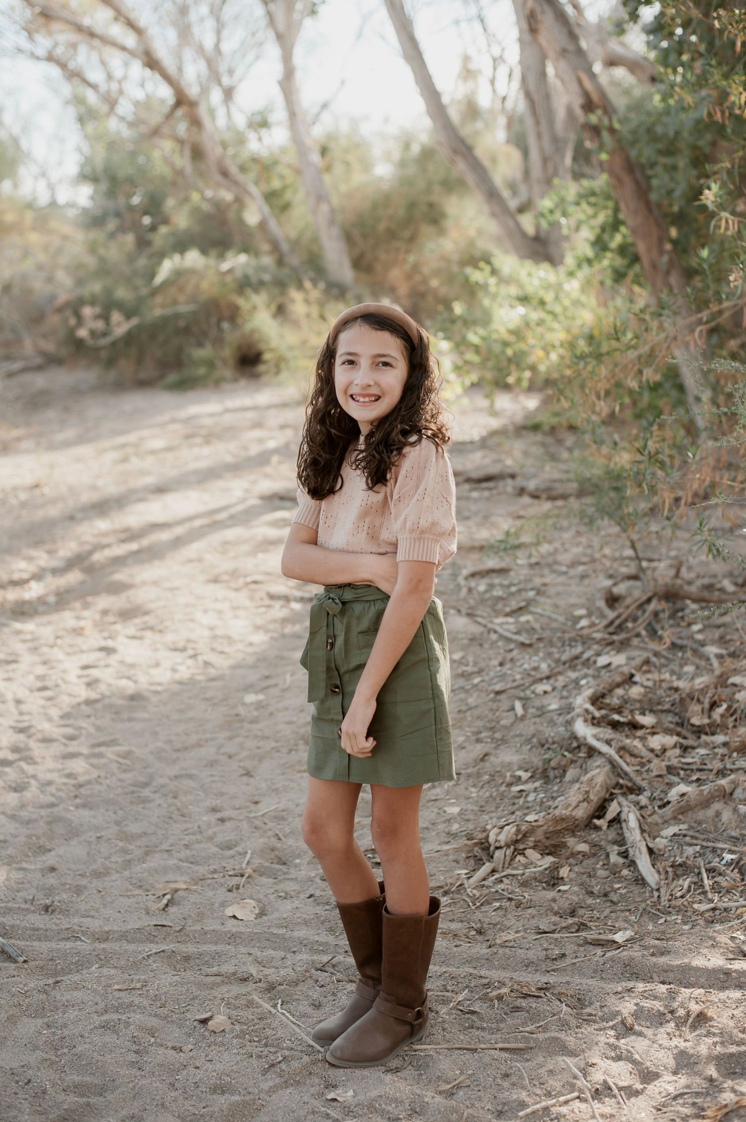 Someone in an olive green romper and brown boots poses playfully on a dirt path surrounded by trees.
