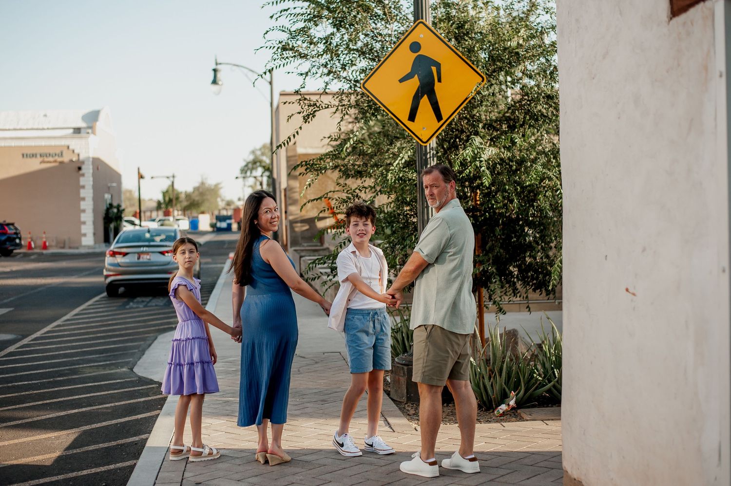 Family of four holding hands on a sunny sidewalk near a pedestrian crossing sign in a urban setting.