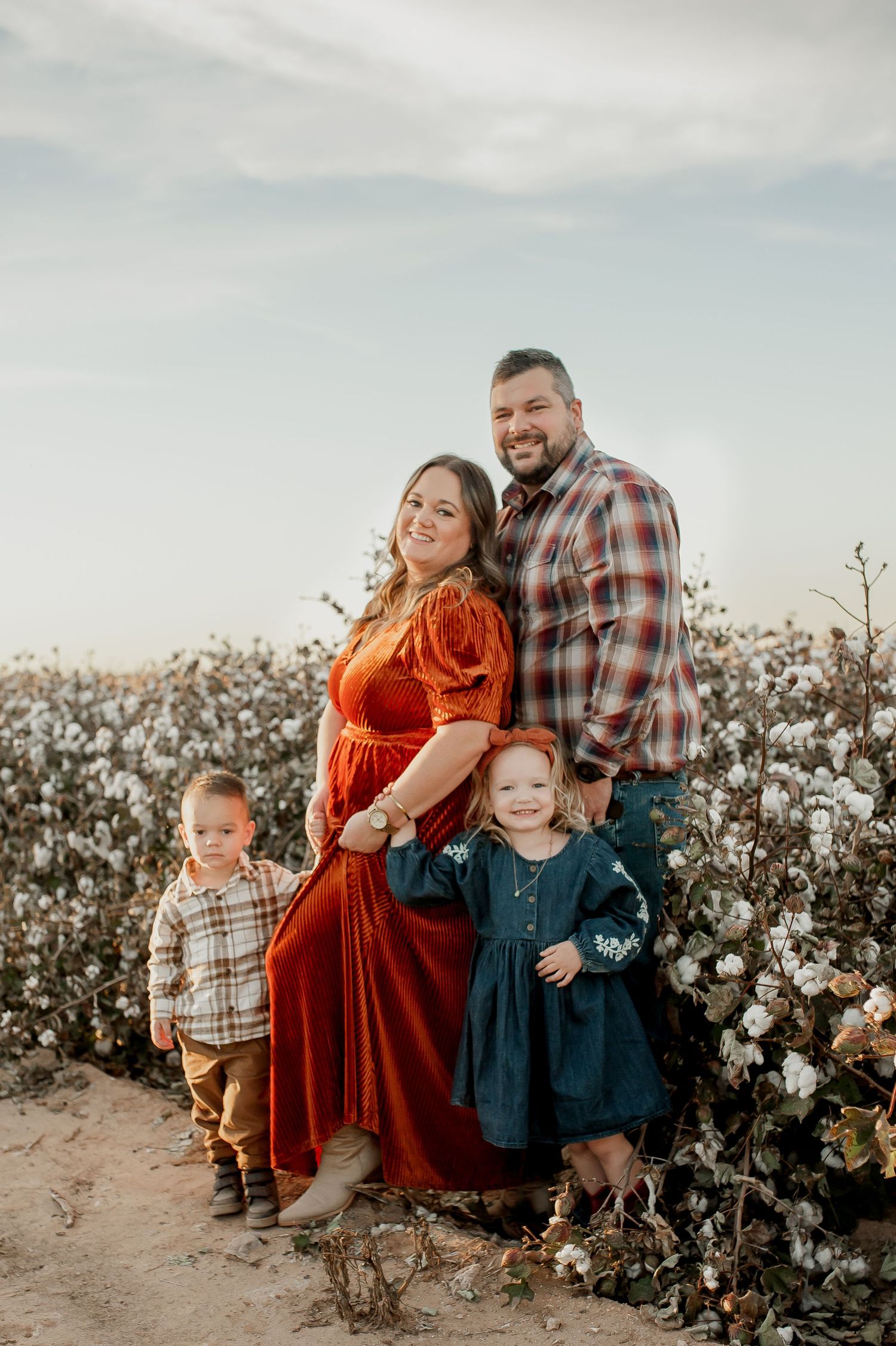 Family of four posing in a cotton field, parents in plaid and red dress with two young children smiling outdoors.