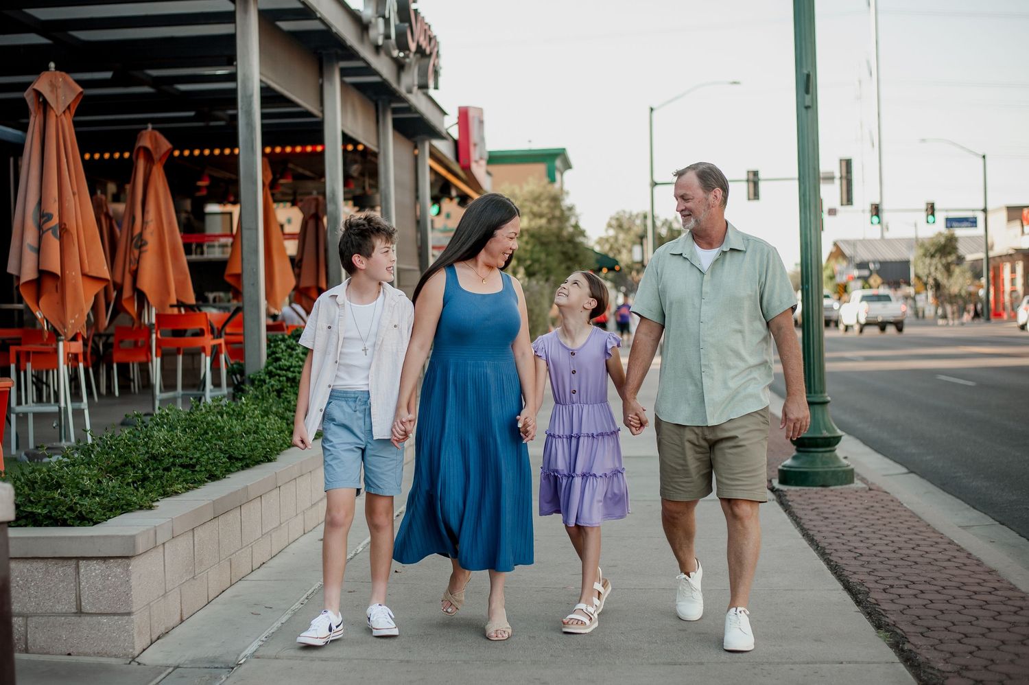 Family of four walking hand-in-hand on a sunny sidewalk past a restaurant patio in a vibrant downtown area.