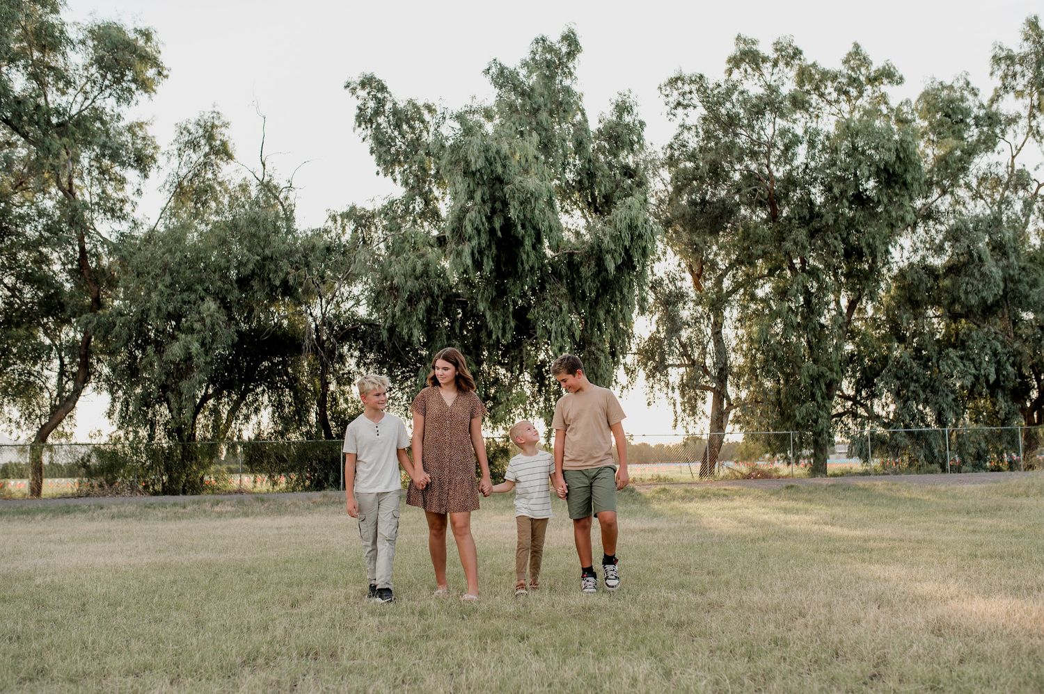 Family of four walking together in a green park surrounded by tall trees during golden hour.