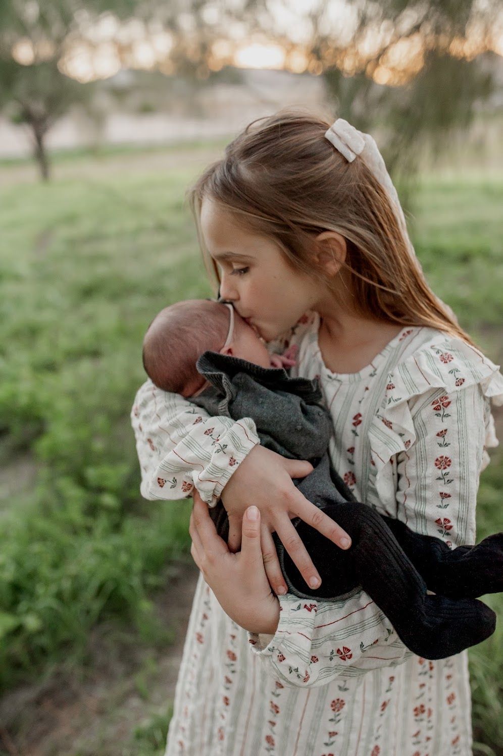 Young girl in floral dress kisses newborn baby she holds gently outdoors in a green field at sunset.