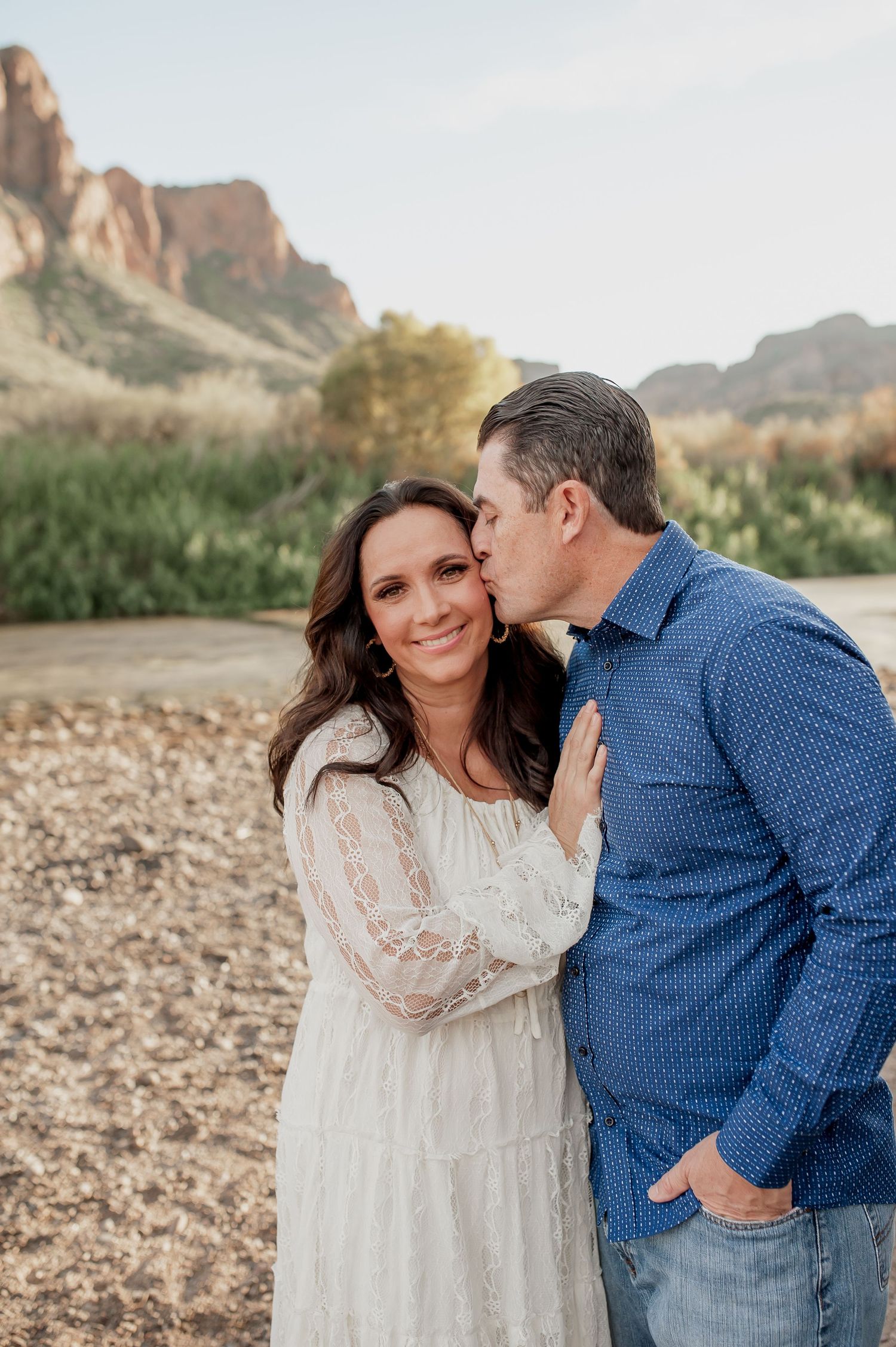 A couple shares romantic moments against a scenic desert mountain backdrop in casual attire.