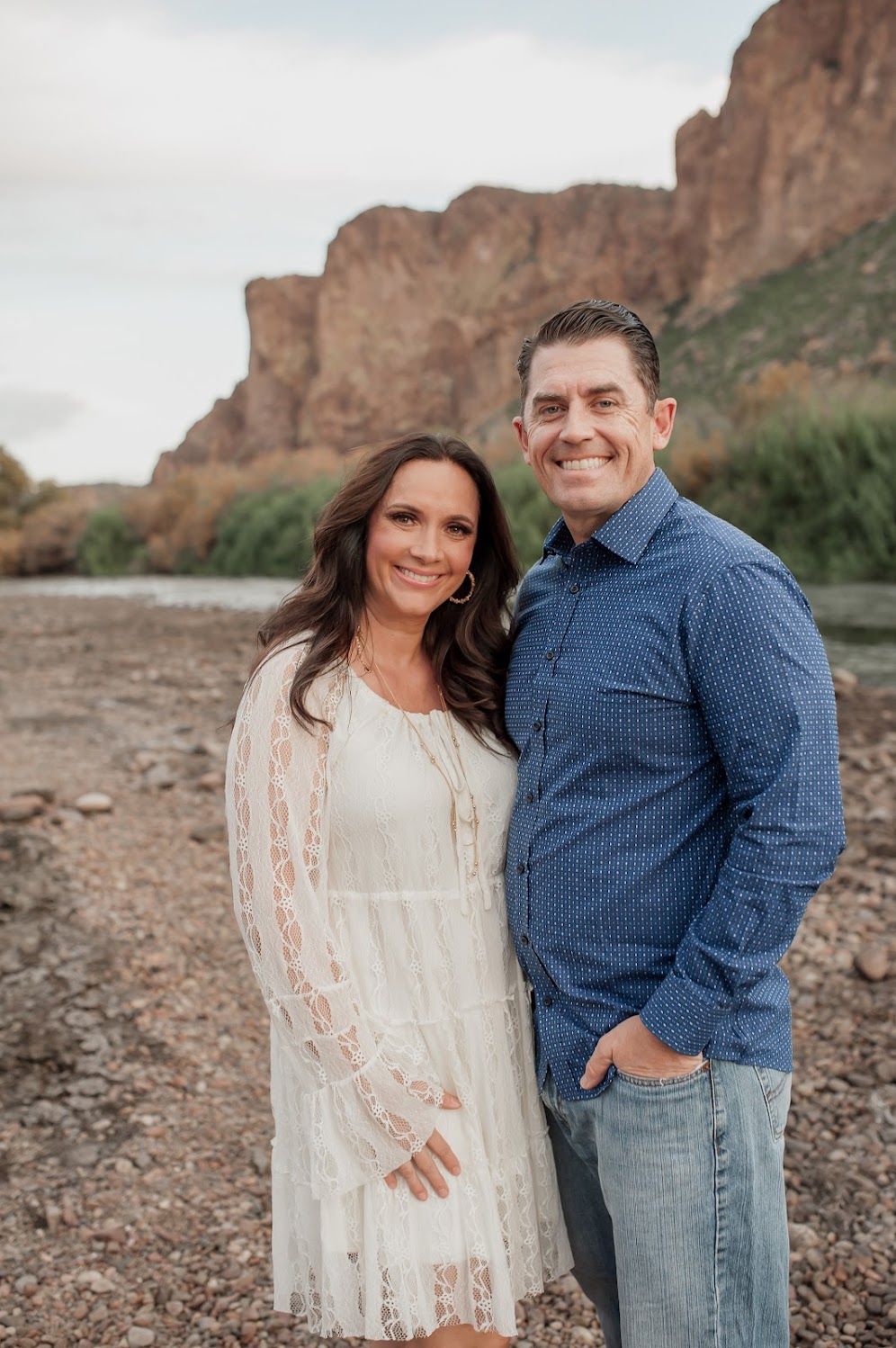 Couple smiling outdoors near rocky desert landscape and river, woman in white lace dress, man in blue shirt and jeans.