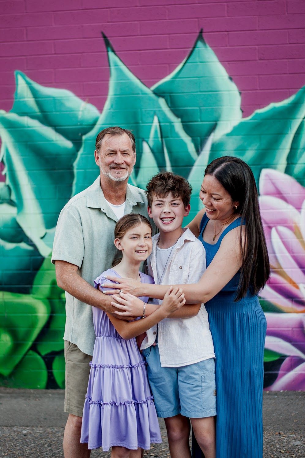 Family of four smiling together in front of a colorful floral mural with pink and green succulents.