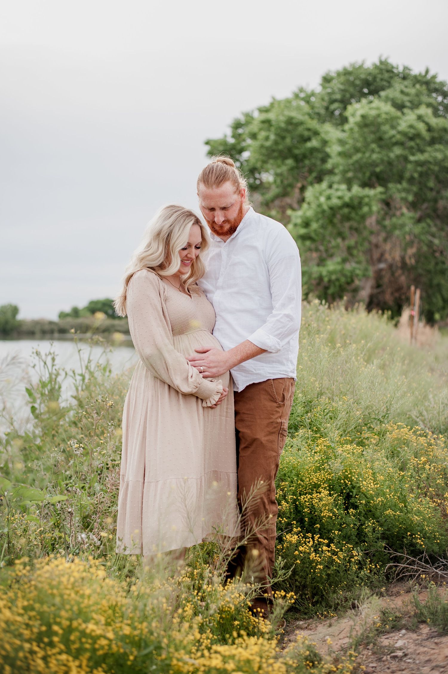 Couple shares intimate moment in a field of wildflowers by a lake during maternity photoshoot.