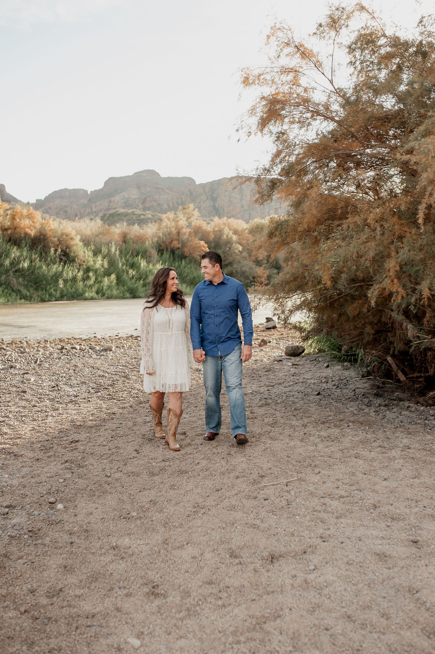 Couple walking along a rocky desert trail with mountains and autumn foliage in the background.