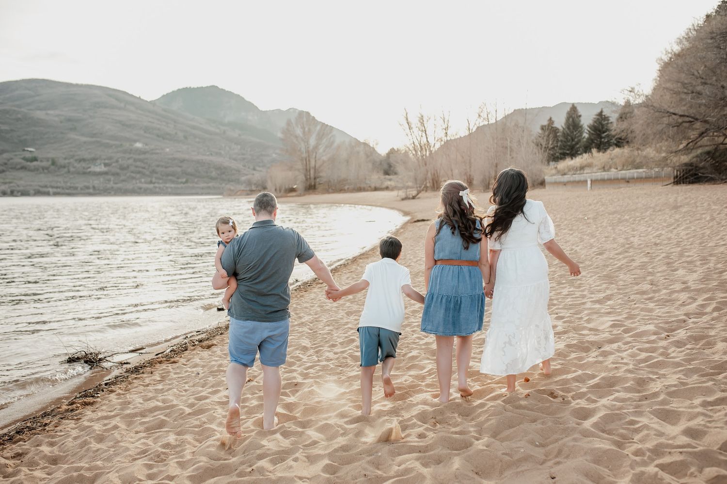 Family of five walking hand-in-hand along a sandy beach shore with mountains and a lake in the background.
