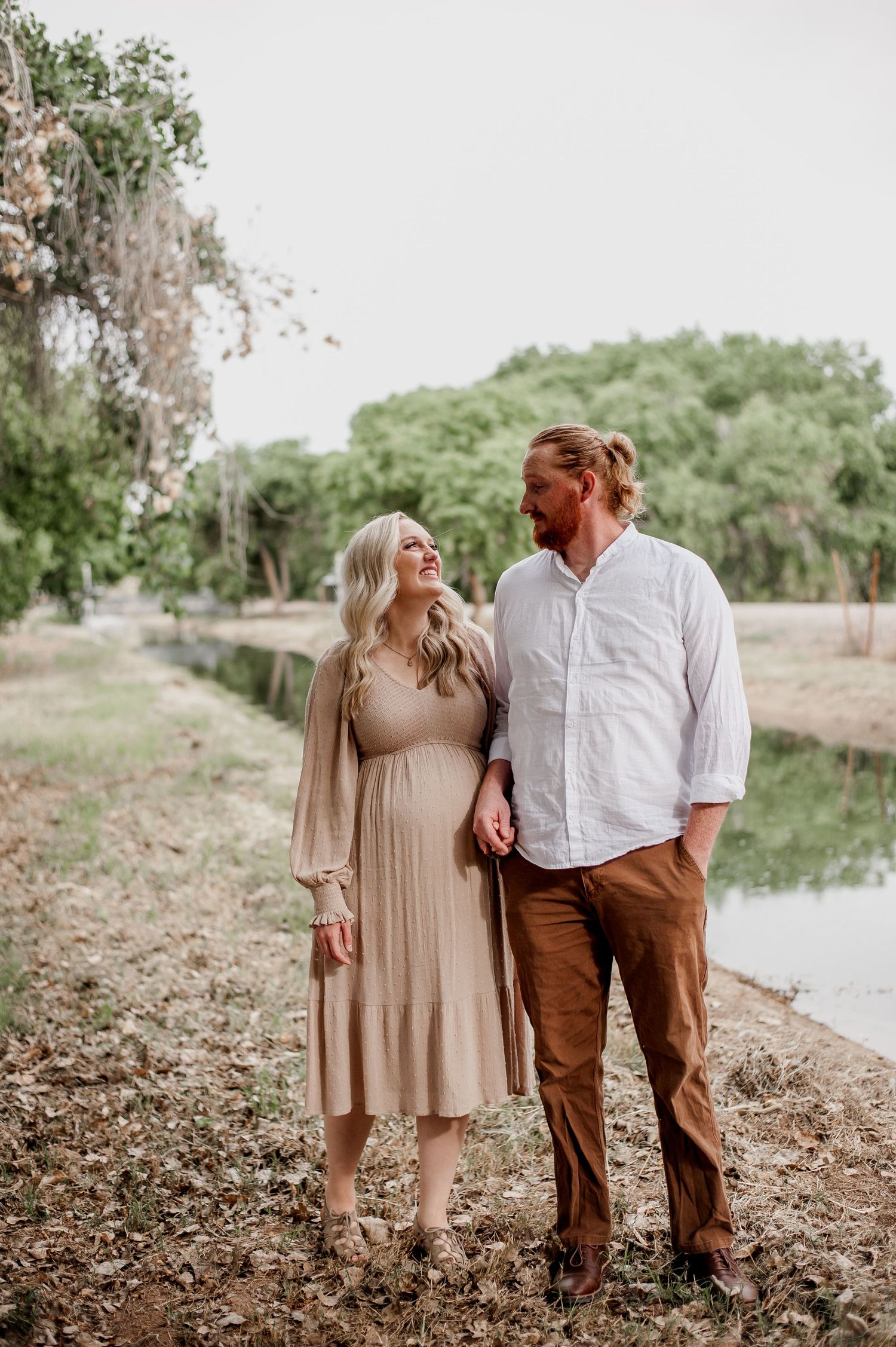 A couple takes romantic photos by a lake during their maternity session in a natural outdoor setting.