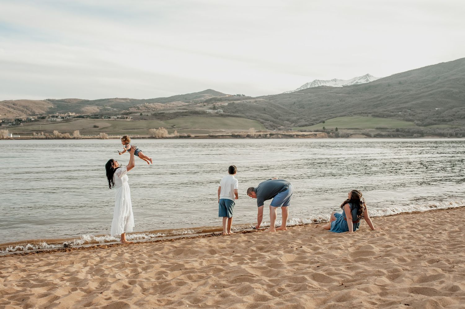 Family enjoying a beach day with mountains in background, parents playing with baby while children explore the shoreline.