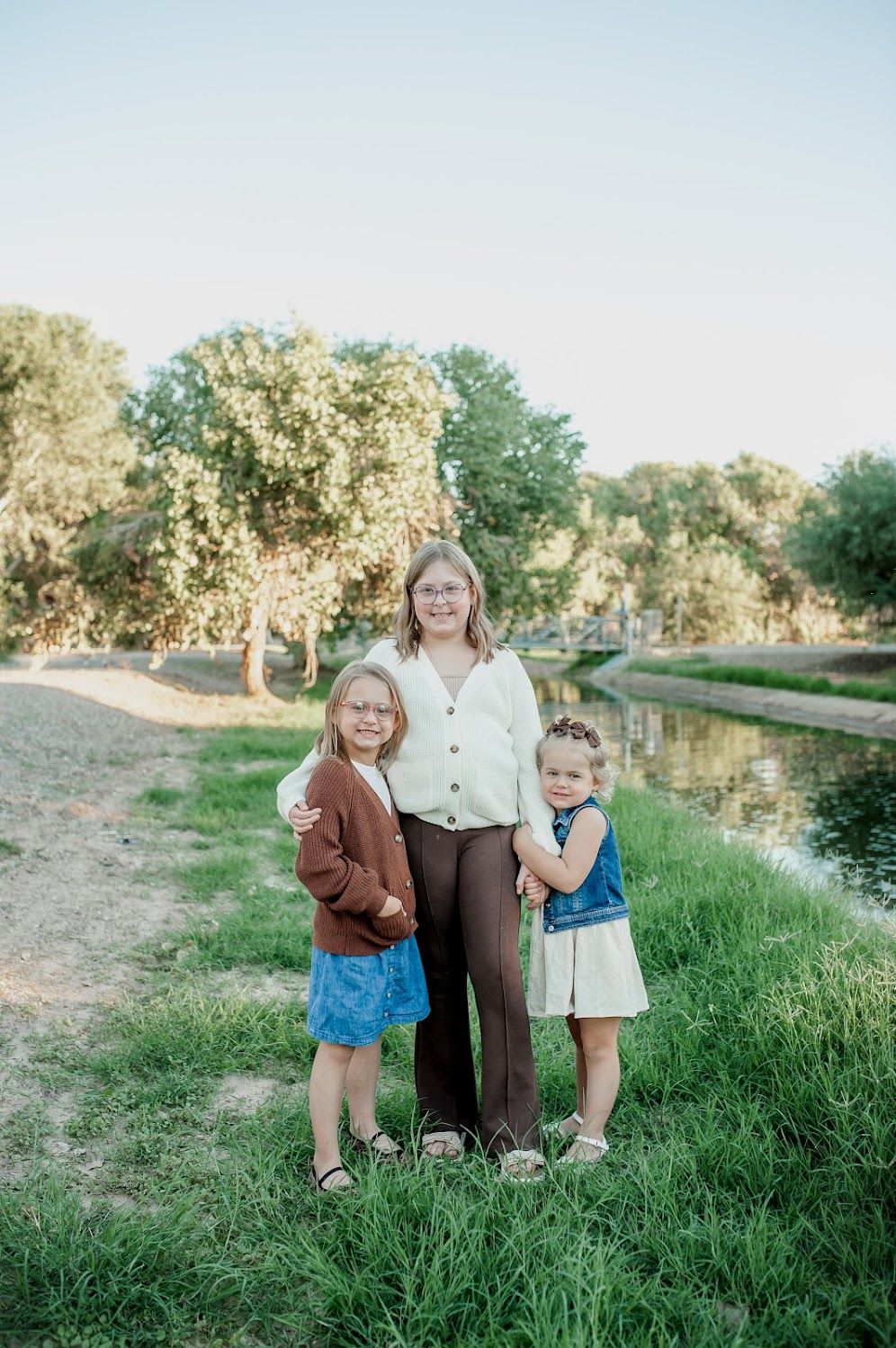 A woman and two young girls pose together on a grassy riverbank surrounded by lush green trees.