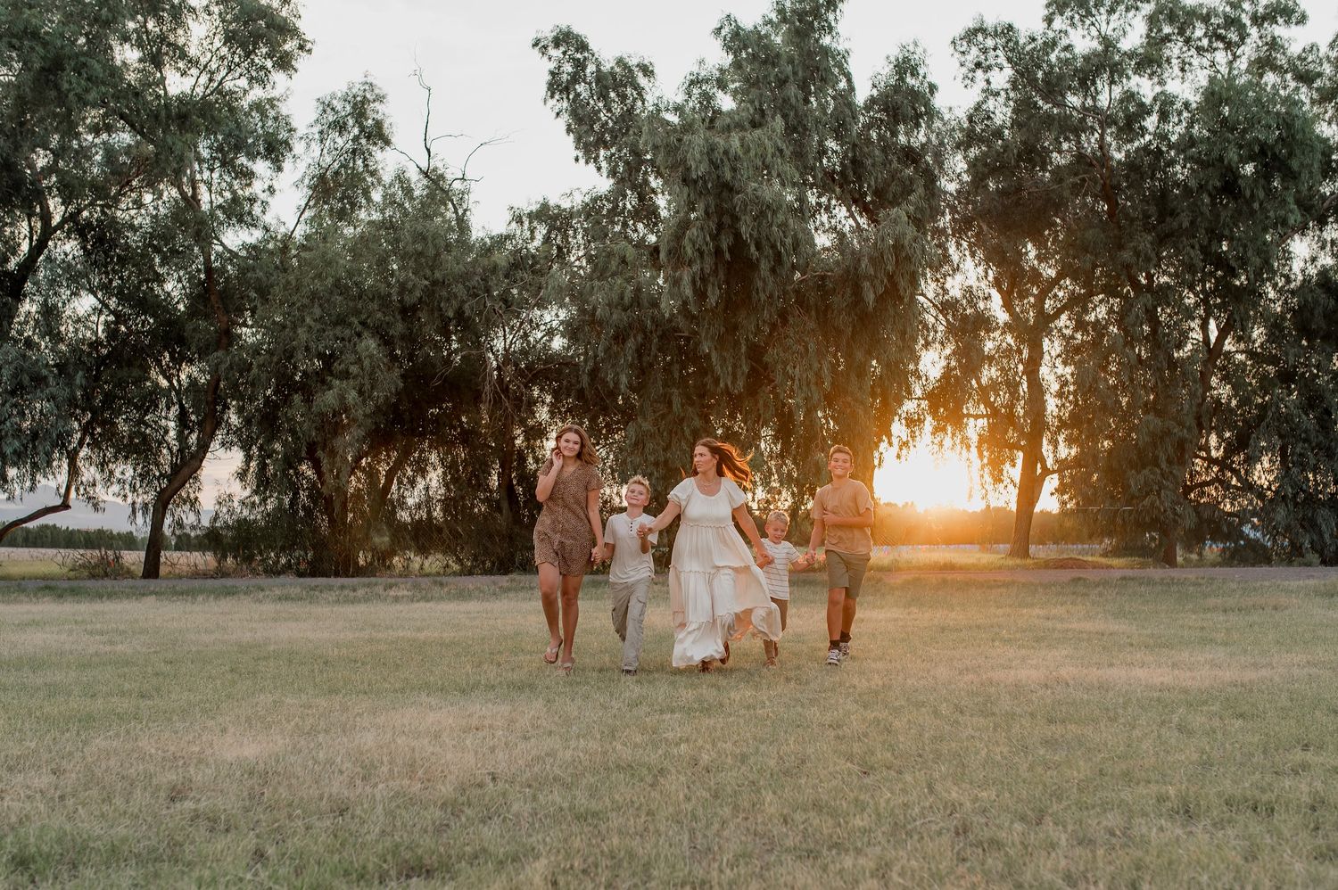 A bridal party walks joyfully through a sunlit field with golden sunset and lush green trees in the background.