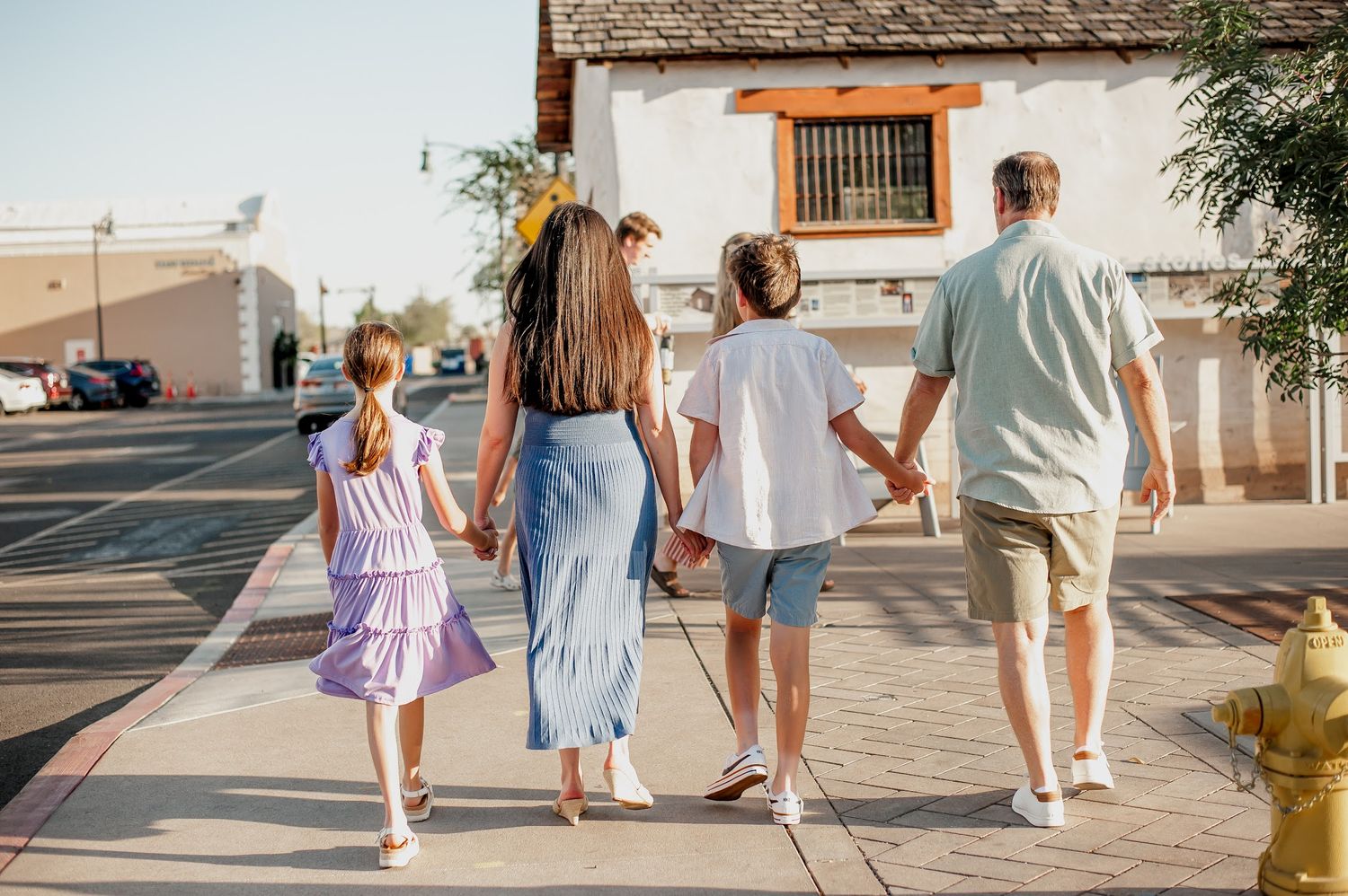 Family of four walking hand-in-hand down a sunny sidewalk in a small town, viewed from behind.