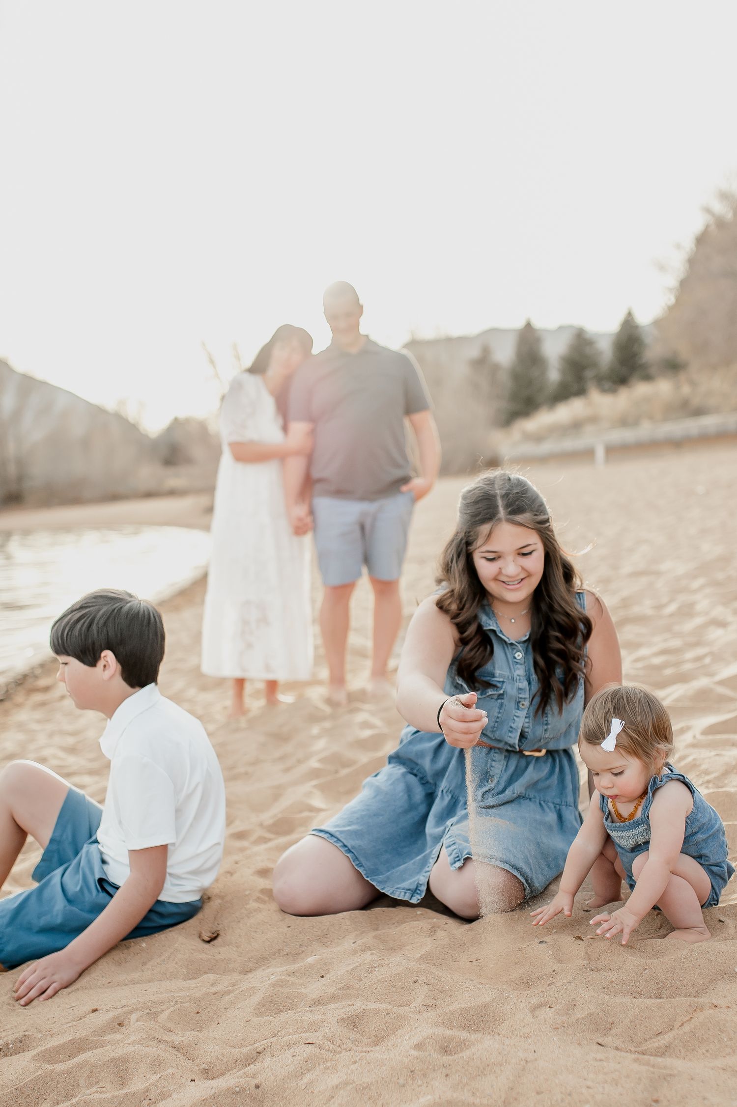 Family beach portrait with two children playing in sand while parents embrace in the background on a sunny day.