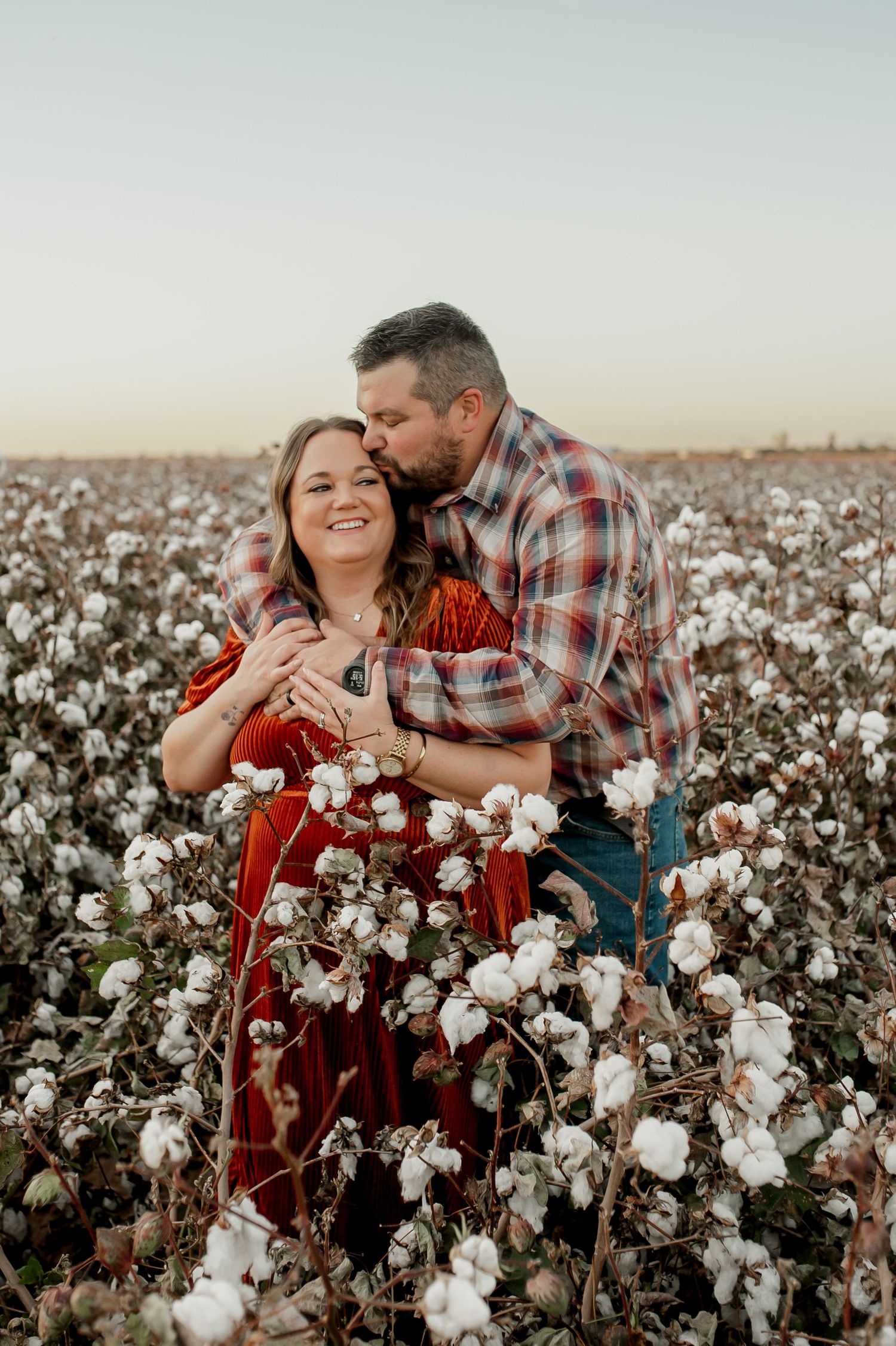Couple embracing in a cotton field at golden hour, woman in red dress, man in plaid shirt kissing her cheek.