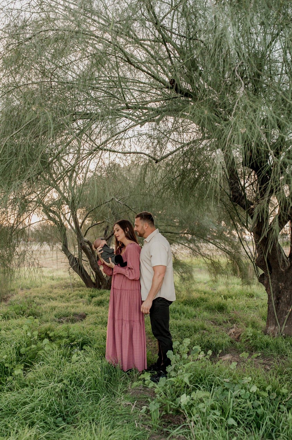 Couple stands under a willow tree in lush greenery, woman in pink dress, man in white shirt, holding a baby.