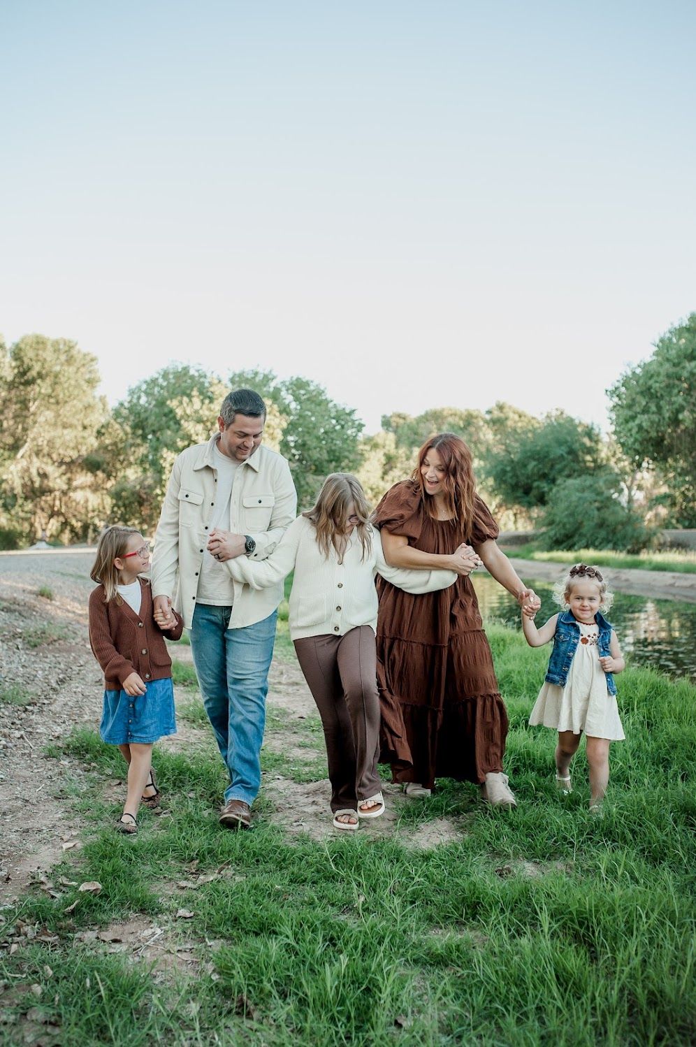 A happy family of five walks together on a grassy path surrounded by trees during a warm outdoor photo session.