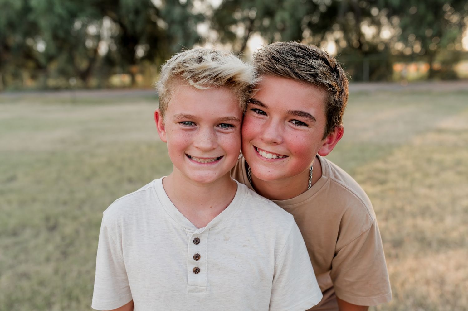 Two smiling boys pose together outdoors in a sunny park, wearing casual shirts in a warm, candid portrait.