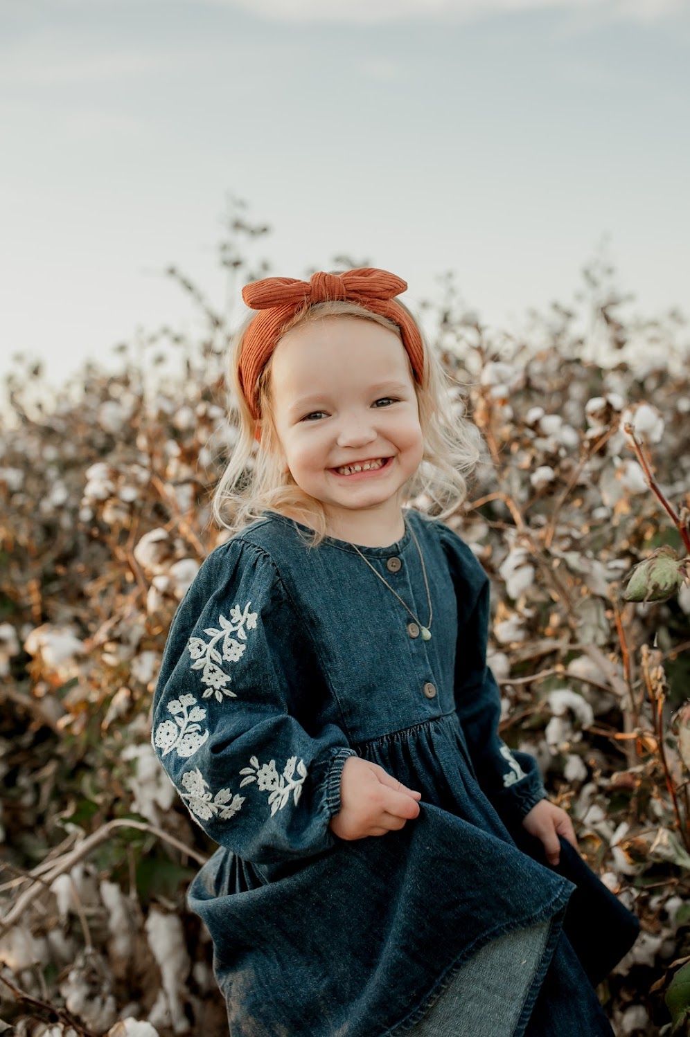 Smiling toddler girl in embroidered denim dress and rust bow headband standing in a cotton field.