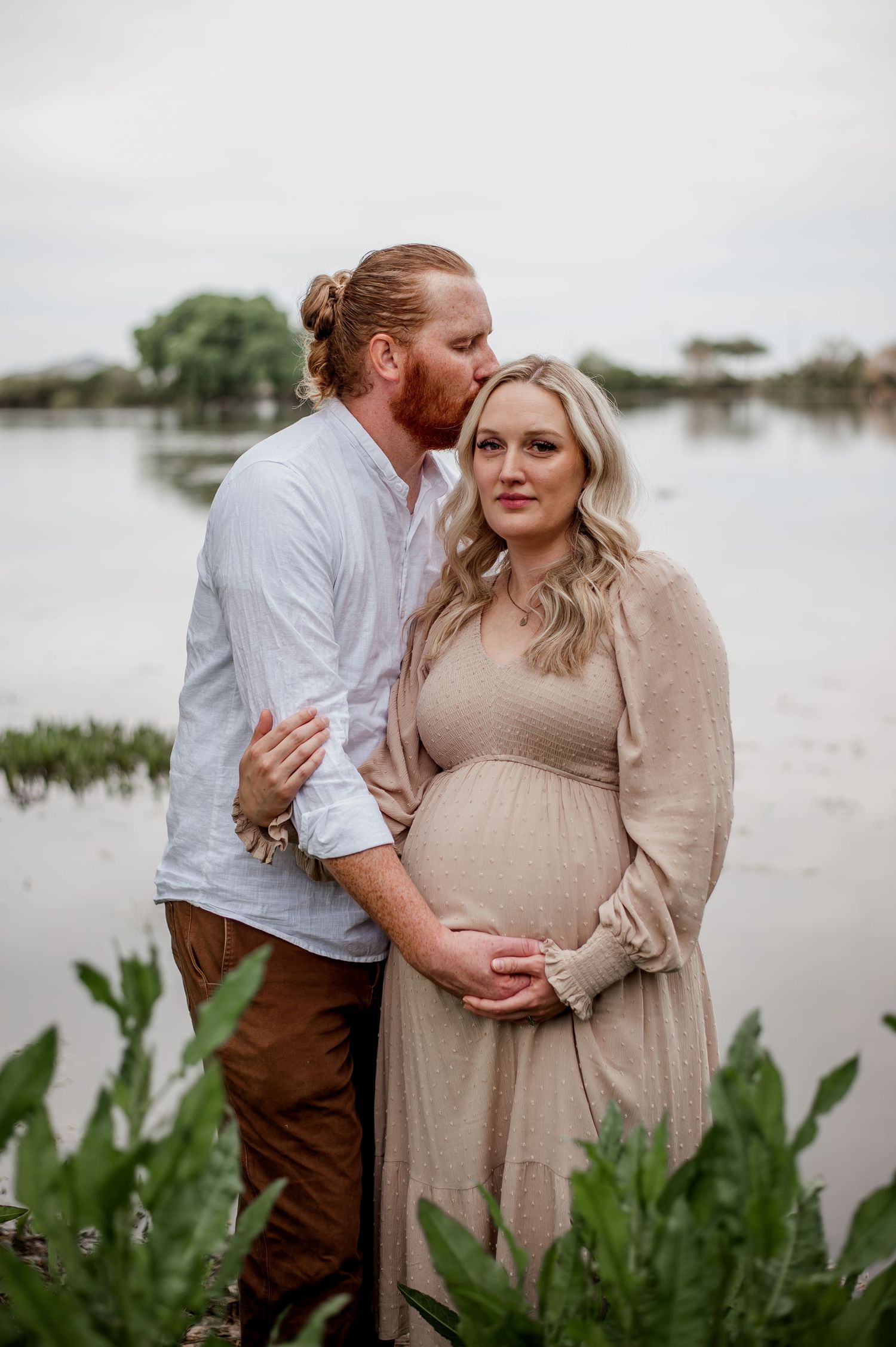 A romantic maternity photoshoot by a lake with lush greenery and water in the background.