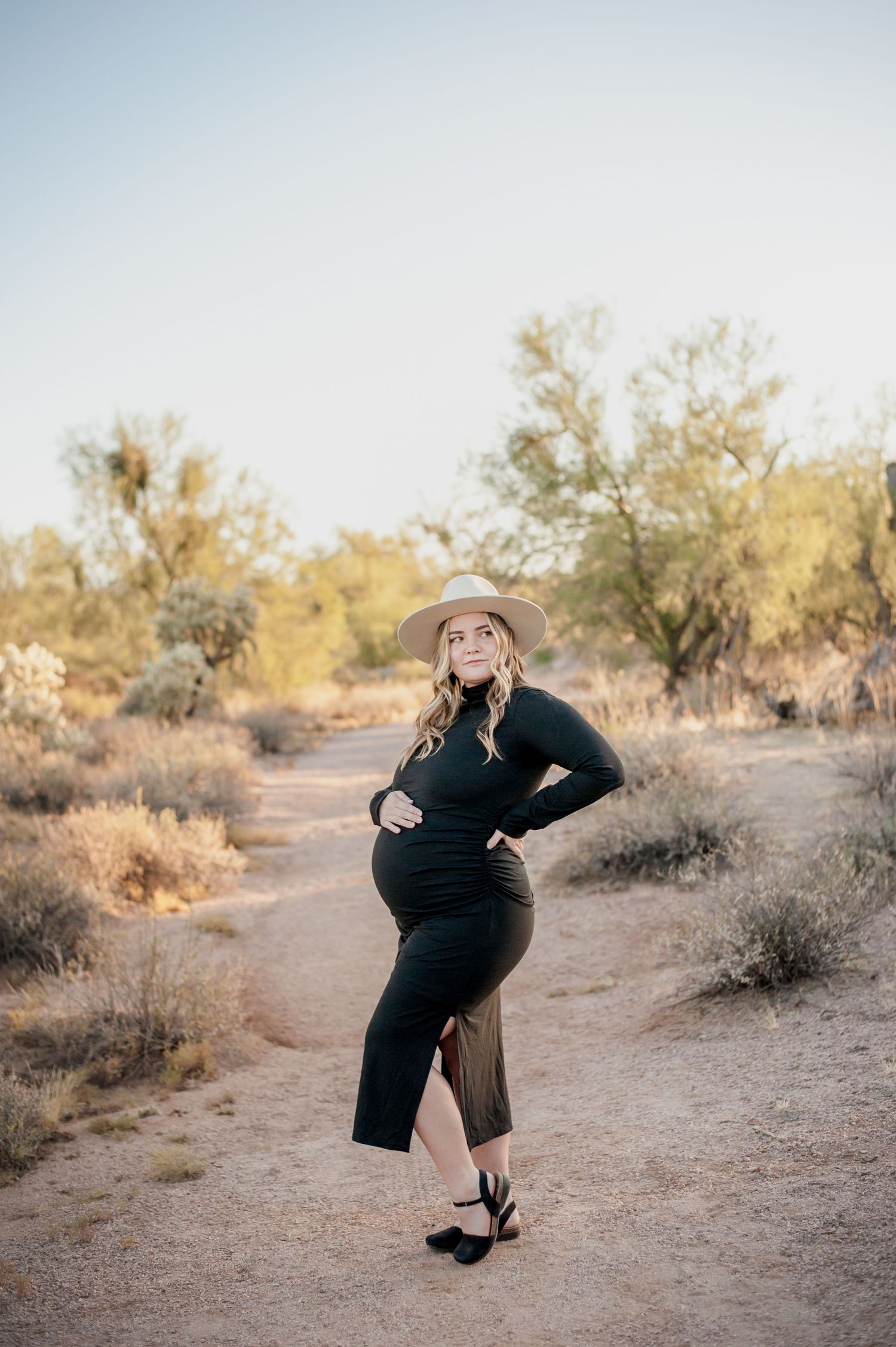 A woman in black dress and hat dances playfully in a desert setting with desert brush in the background.