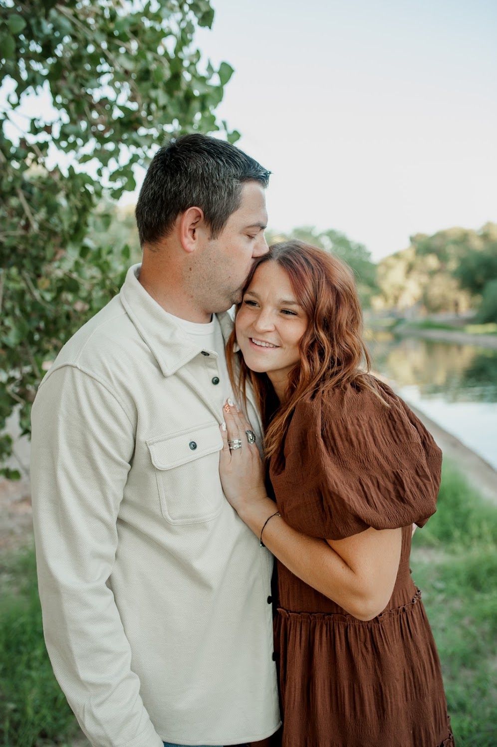A man in a cream shirt kisses a smiling woman in a brown dress outdoors near a serene waterway.