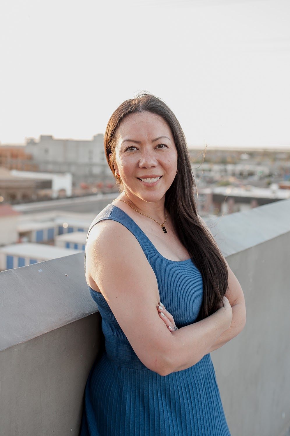 Smiling woman with long dark hair in a blue dress, arms crossed on a rooftop with city views.